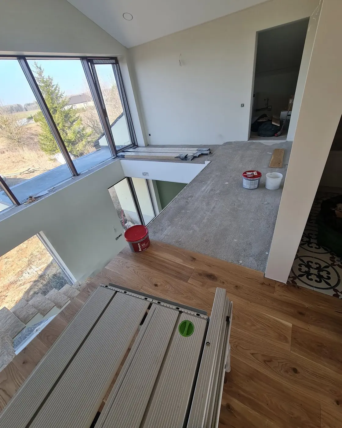 A room at construction stage with panoramic windows: oak boards already laid on one side, bare concrete substrate on the other