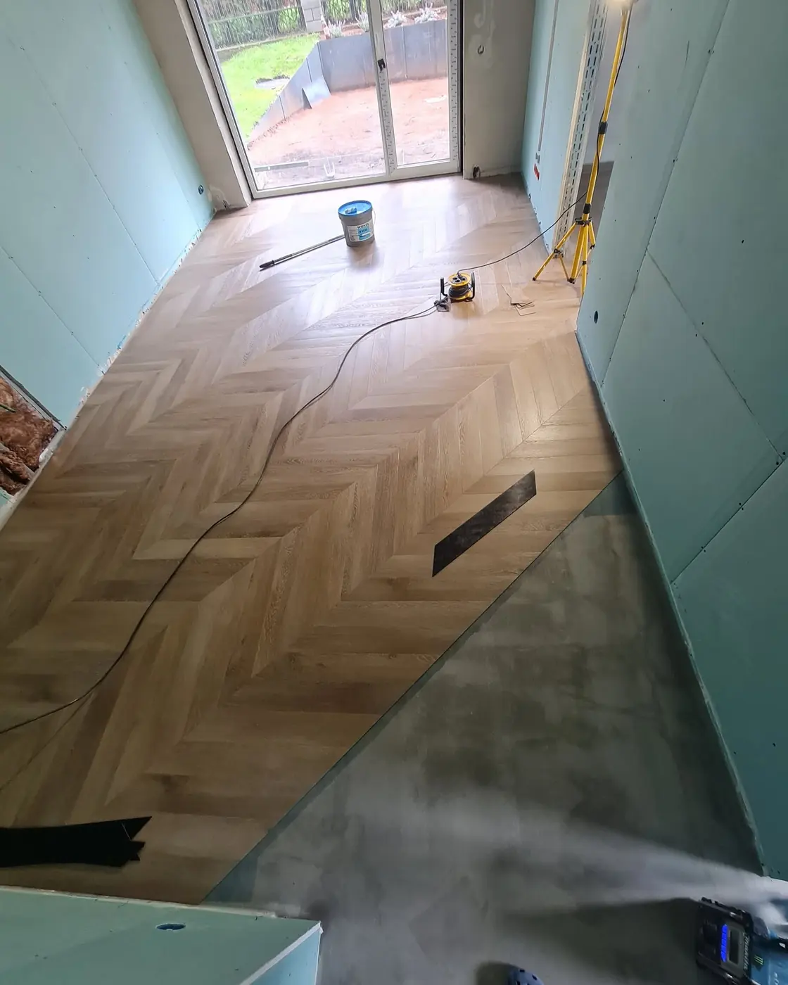 Half the room covered in oak chevron, the rest still bare concrete, with a work light and a bucket of adhesive beside it