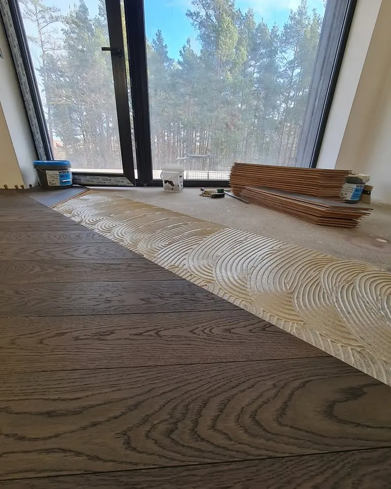 A band of combed adhesive at the edge of the laid dark oak boards, with angle-cut chevron boards stacked by the window